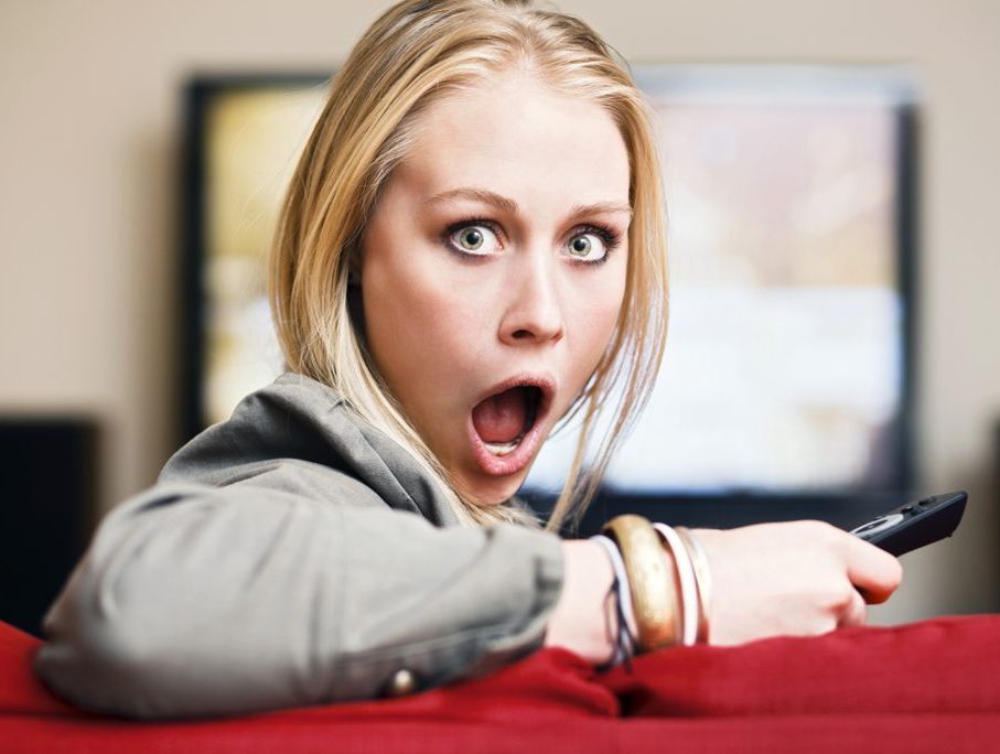 Blonde watching TV and holding remote control looks round, horrified.